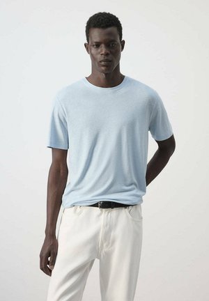 Hombre de pie frente a un fondo liso, vestido con una camisa de manga corta azul claro y pantalones blancos con cinturón negro.