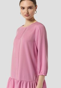 Rosafarbenes langärmliges Kleid mit Rundhalsausschnitt, elastischen Bündchen, einem gestuften Volantsaum und subtilen Falten vorne, aus leichtem Stoff gefertigt.