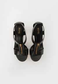 Paire de sandales en cuir noir à bout ouvert avec fermetures éclair dorées à l’avant et brides à la cheville, posées à plat sur fond blanc.