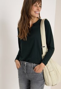 Un haut à manches longues vert foncé avec un col henley, associé à un jean en denim gris. Un sac à épaule de couleur crème est porté en bandoulière.