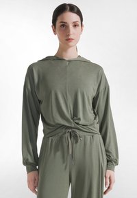 Kobieta ubrana w przygaszony zielony, z kapturem, długorękawowy top z przednim skrętem oraz dopasowane spodnie ze ściągaczem, stojąca na jednolitym tle.
