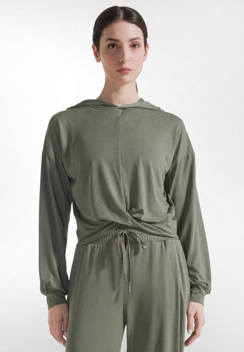 Kobieta ubrana w przygaszony zielony, z kapturem, długorękawowy top z przednim skrętem oraz dopasowane spodnie ze ściągaczem, stojąca na jednolitym tle.