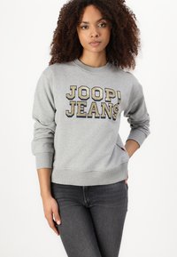 Felpa grigia con "JOOP! JEANS" in grandi lettere dorate e blu navy. Presenta polsini e orlo a coste. Indossata con jeans scuri.