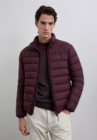 Chaqueta de abrigo burdeos con diseño de acolchado horizontal, cuello alto y un logo sutil. Llevada sobre un suéter gris oscuro, combinada con pantalones claros.