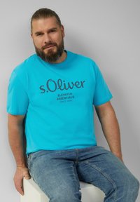 Mėlynas medvilnės marškinėliai su trumpomis rankovėmis ir apvalia iškirpte, featuring tamsiai pilką "s.Oliver" logotipą ir "Elevated Essentials" tekstą.