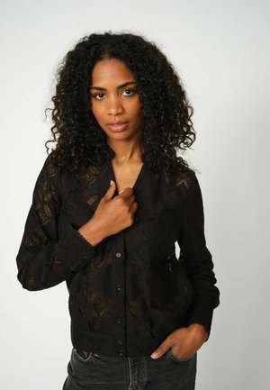 Black lace button-up blouse with long sleeves, floral pattern, delicate scalloped cuffs, and textured finish. Worn over dark jeans.