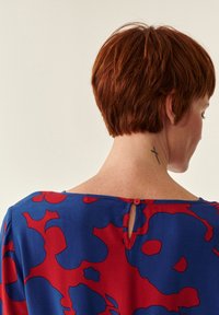 Bluse med blå og rød blomsterprint, der har en lille knaplukning i ryggen og en rund halsudskæring, lavet af letvægtsstof. Synlig nakketatovering.
