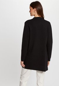 Schwarzer langärmeliger Cardigan in gerader Form, aus weichem Material gefertigt, mit Kragen und ohne sichtbare Verschlüsse, über hellen Hosen getragen.