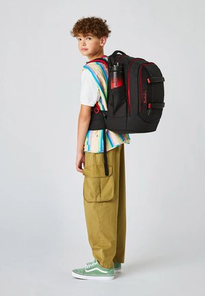 Ragazzo con zaino nero e rosso con bottiglia d'acqua, camicia a righe multicolore, pantaloni cargo color kaki e scarpe da ginnastica verdi, visto di lato.