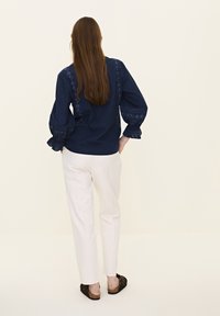 Marineblaue Bluse mit floralem Stickerei entlang der Ärmel und am Rücken, kombiniert mit weißen Hosen und dunklen, flachen Sandalen. Glatte Textur.
