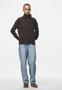 Brun stickad turtlenecktröja med en avslappnad passform, kombinerad med ljusblå raka jeans. Modellen står med händerna på höfterna.