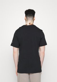 Muž s krátkými kudrnatými vlasy a vitiligem na krku, oblečený v jednoduchém černém tričku s krátkým rukávem a béžových kalhotách, stojící proti bílému pozadí.