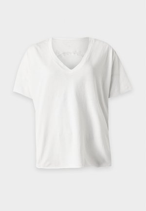 Kvinde stående iført en hvid V-hals T-shirt, sorte bukser med vide ben og hvide sneakers mod en ensartet lys baggrund.