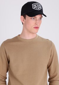Gorra de malla negra con el logo blanco "DEUS." combinada con una camiseta de manga larga beige que presenta un cuello redondo y una textura suave.