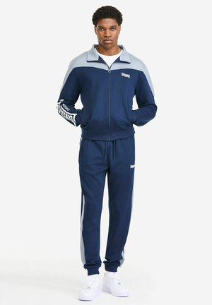 Granatowy dres z jasnoniebieskimi akcentami; kurtka na zamek błyskawiczny, ściągacz przy kołnierzu i mankietach; spodnie typu jogger z detalami logo i bocznymi paskami.