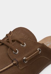 Chaussure à enfiler en daim marron avec lacets en cuir passés dans des œillets argentés, coutures apparentes et design à talon ouvert.