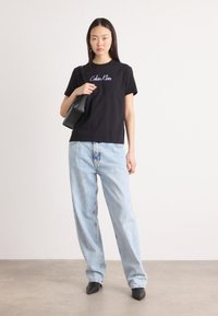 Crna pamučna majica s ljubičastim logom "Calvin Klein", uparen sa svijetloplavim širokim trapericama i crnim cipelama s oštrim vrhom. Crna torba.