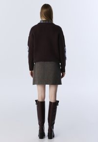 Vrouw staat, gekleed in een donkerbruine trui met lichtblauwe vlekken, een houndstooth rok en hoge donkerbruine laarzen tegen een effen achtergrond.