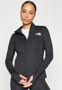 Chaqueta negra con cremallera hecha de tela elástica, con cuello alto, agujeros para los pulgares en las mangas y un logo blanco en el pecho izquierdo.