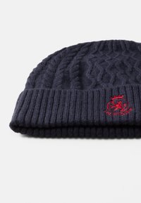 Navyblauwe gebreide beanie met een gestructureerd kabelpatroon en een omgevouwen ribkraag. Bevat een rood geborduurd logo met een leeuw en kroon.