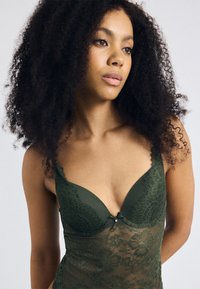 Lingerie in pizzo verde oliva con scollatura profonda, motivi floreali, bordi ondulati e un piccolo fiocco al centro.