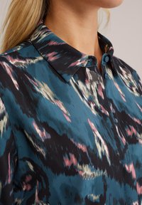 Blusa in satin con un motivo astratto nero, verde teal e rosa, dotata di colletto classico, chiusura a bottoni e una consistenza morbida e liscia.