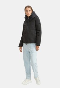 Giacca nera isolata con zip frontale e grande cappuccio, indossata sopra jeans azzurri chiari e sneakers bianche. Tessuto liscio con dettagli minimi.