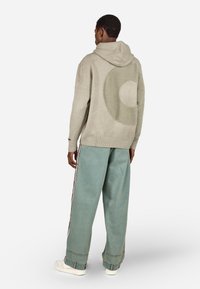 Világosszürke kapucnis pulóver, melynek hátoldalán texturált kör alakú minta található, laza világoskék nadrággal és fehér cipővel párosítva.