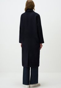 Marineblauwe jas met een gestructureerde kraag en lange mouwen, met een rechte snit en zijsplitten, gecombineerd met wijde denim jeans en witte sneakers.
