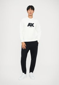 Fehér pulóver fekete "AX" logóval, hozzá illő sötétkék melegítőnadrággal és fehér sportcipővel. Az öltözet laza, kényelmes fazonú.