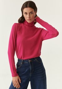 Maglione a collo alto aderente in tessuto a coste rosa brillante, abbinato a jeans in denim blu scuro a vita alta. Maniche lunghe e texture minimale.