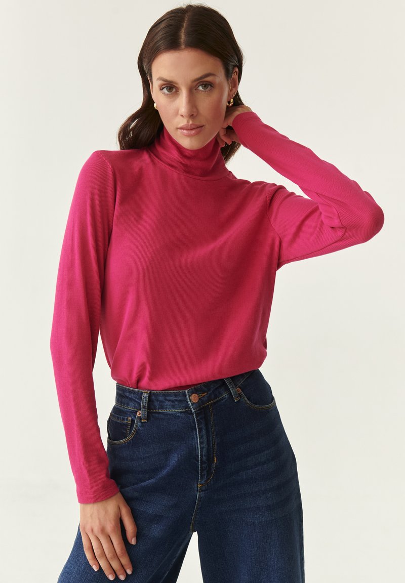 Maglione a collo alto aderente in tessuto a coste rosa brillante, abbinato a jeans in denim blu scuro a vita alta. Maniche lunghe e texture minimale.