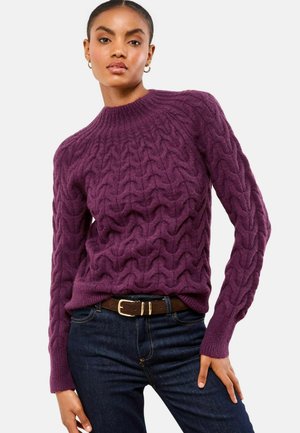 Purple cable-knit sweater with a high neckline and textured pattern. Paired with dark jeans and a brown belt accentuating the waist.