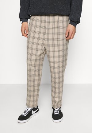 Mężczyzna ubrany w beżowe spodnie w kratę o prostych nogawkach oraz czarno-białe sneakersy, stojący na tle jednolitego białego tła.