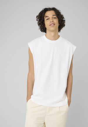 Jeune personne aux cheveux bouclés portant un débardeur blanc et un pantalon beige clair, debout les mains dans les poches devant un fond gris.