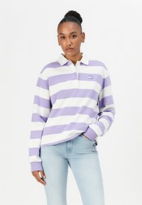 Žena s visokim punđom koja nosi dug rukav, ljubičasto-bijelu prugastu polo majicu i svijetloplave traperice, stoji naslonjena na bijelu pozadinu.