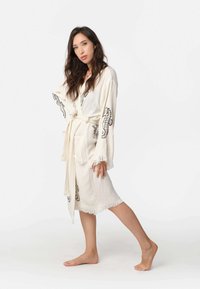 Svetlo béžový textúrovaný župan s čiernym motívom morského koníka, zaviazaný v páse. Má dlhé rukávy a strapcové detaily na spodnom leme.