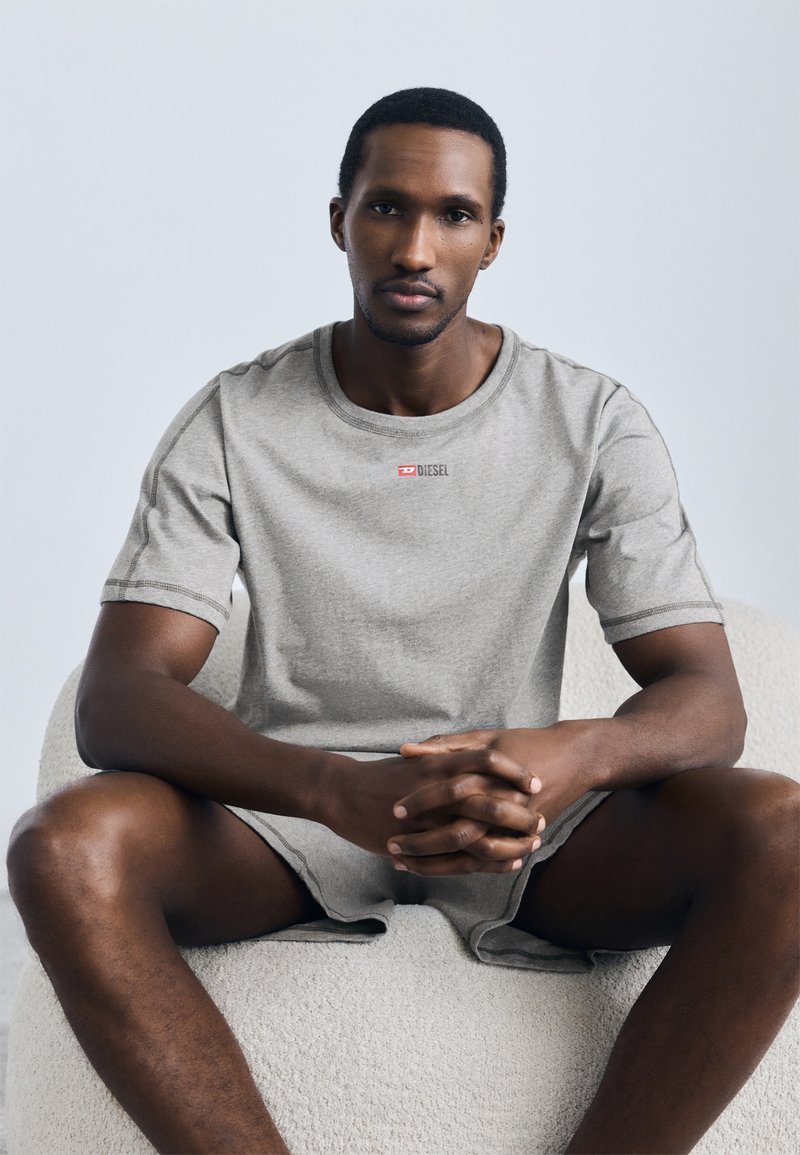Γκρι βαμβακερό t-shirt με κοντά μανίκια, διακοσμημένο με κόκκινο λογότυπο Diesel στο στήθος. Το μοντέλο κάθεται με σταυρωμένα πόδια σε μια καρέκλα με υφή.