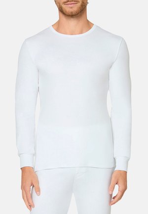 Mann trägt ein schlichtes, eng anliegendes weißes Langarmshirt und passende weiße Hosen vor neutralem Hintergrund.