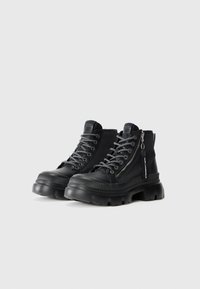 Zwarte enkellaarzen met een chunky zool, gemaakt van textiel en leer. Voorzien van grijze veters en een zijrits met een metalen trekker.