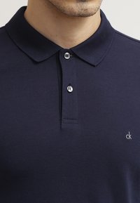 Granatowa koszulka polo z teksturowanej tkaniny, z klasycznym kołnierzykiem, zapięciem na dwa guziki oraz małym haftowanym logo na piersi.