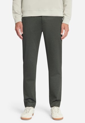 Unterkörper einer Person, die dunkelgraue Slim-Fit-Hosen, off-white Sneaker und ein hellbeiges langärmeliges Oberteil vor einfachem Hintergrund trägt.