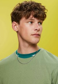 Grøn bomulds t-shirt lagdelt over en tealfarvet skjorte, sølv kæderekæde, kort bølget hår, mod en lys gul baggrund.