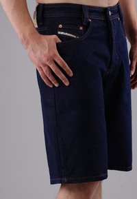 Dark blue denim shorts with a straight cut, featuring copper rivets, contrast stitching, and a front pocket with patterned fabric detailing.