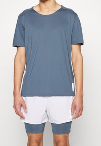 Modrá atletická košile s krátkým rukávem a texturovaným materiálem, v kombinaci s bílými kraťasy. Obsahuje kulatý výstřih a diskrétní branding na lemu.