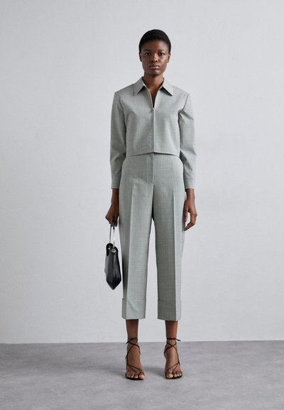 Veste courte en tartan gris avec col pointu, associée à un pantalon taille haute effilé. Le mannequin tient un petit sac à main noir et porte des talons avec des lanières.