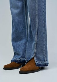 Blue denim jeans with frayed hems slightly covering brown suede pointed-toe boots with low black heels on a gray background.