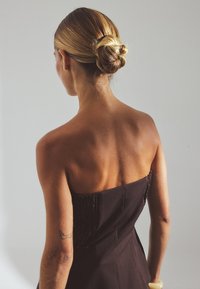 Strapless brown dress with textured bodice, revealing back, and a fitted shape. Hair styled in a low bun; arm features a tattoo.