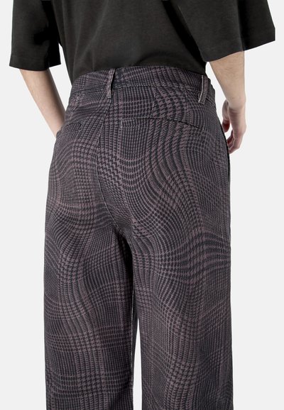 Ansicht von hinten: Person trägt ein schwarzes Oberteil und eine hochtaillierte, weit geschnittene Hose mit einem verzerrten schwarz-lila Karomuster.