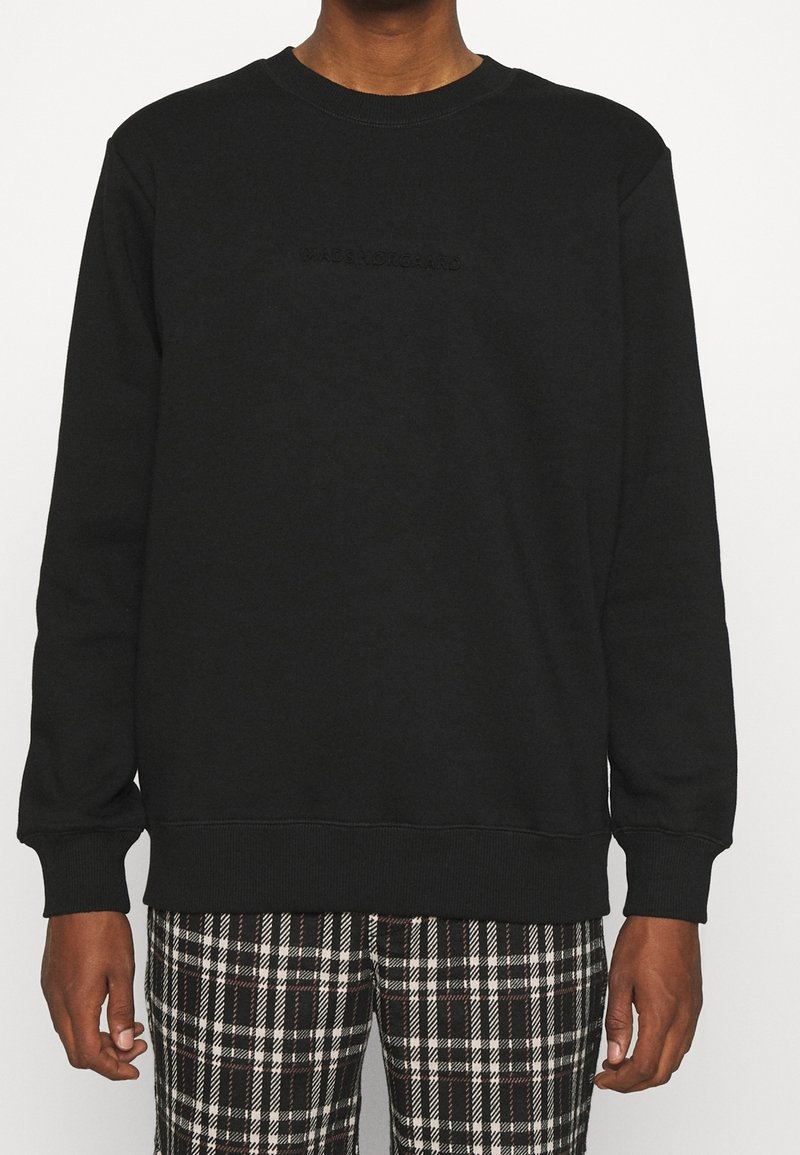 Personne portant un sweat-shirt noir uni à manches longues et un pantalon à carreaux noir et blanc, debout les bras détendus le long du corps.